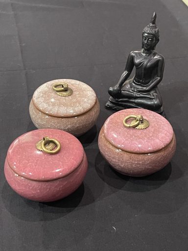 Three decorative jars in pink and cream with a golden lid, beside a small Buddha statue.